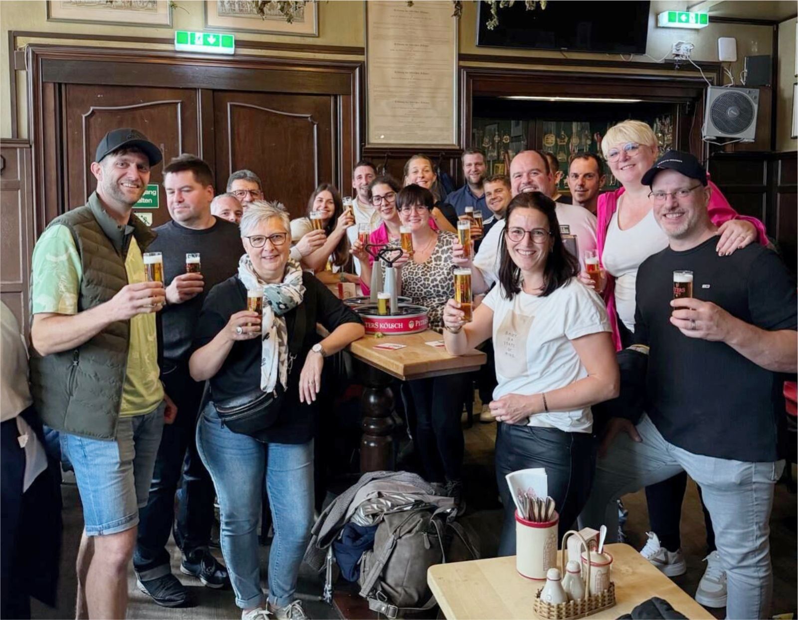 In einer gemütlichen Kneipe mit Holzwänden und Holztischen posiert eine Gruppe lächelnder Menschen mit Getränken in der Hand. Einige stehen, einige sitzen, und alle sehen fröhlich und entspannt aus.